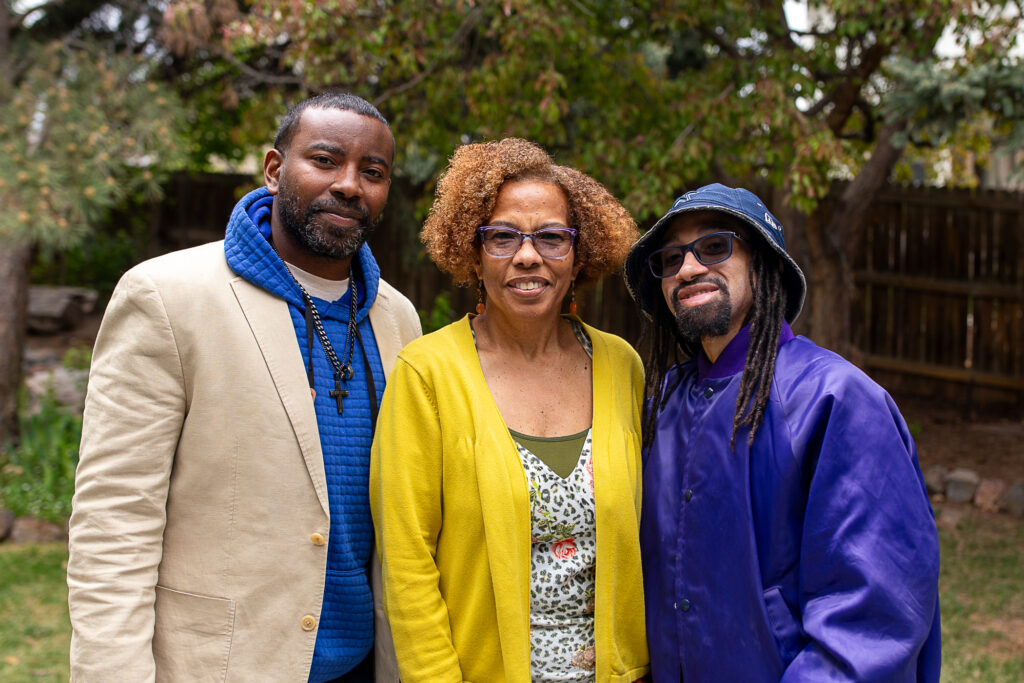 A family of 3 people, tow men and 1 woman, stand outside and pose for a picture, smiling.