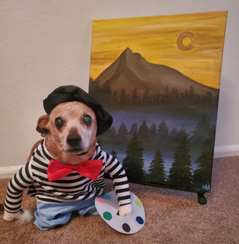 A dog in a painter's outfit and holding a painters pallete stands beside a colorful canvas with a painting of a mountain.