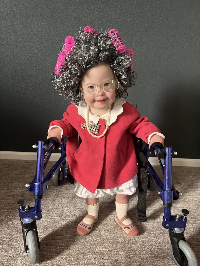 A little girl in a red dress, grey wig with rollers, and glasses. She is standing with a blue walking aid.