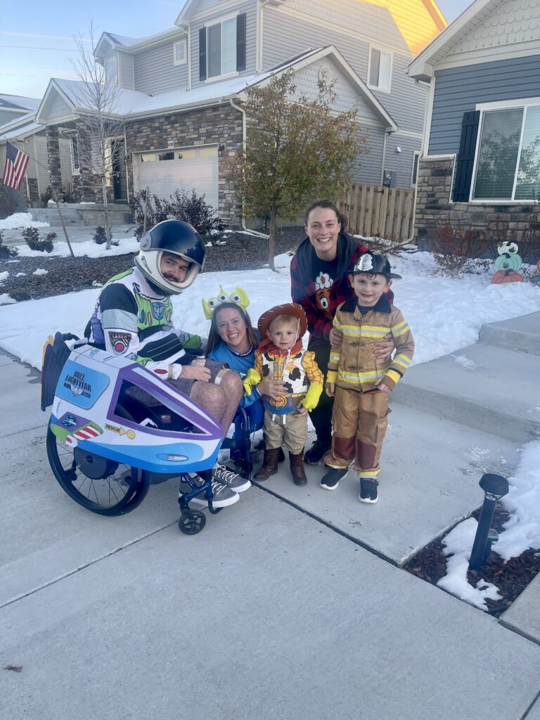 A family in costumes smiles together dressed as toy story characters. A man in a wheelchair is dressed as buzz lightyear, with a cardboard spaceship. A woman is dressed as an alien. A young boy is dressed as Woody. An older boy is dressed as a firefighter.