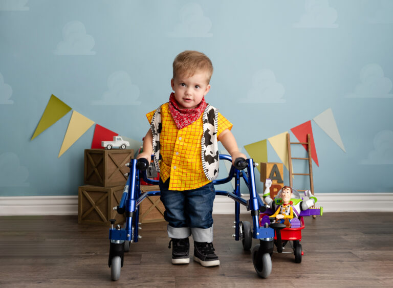 A young boy dressed as woody from toy story standing wit a walking aid. He is surrounded by colorful toys.