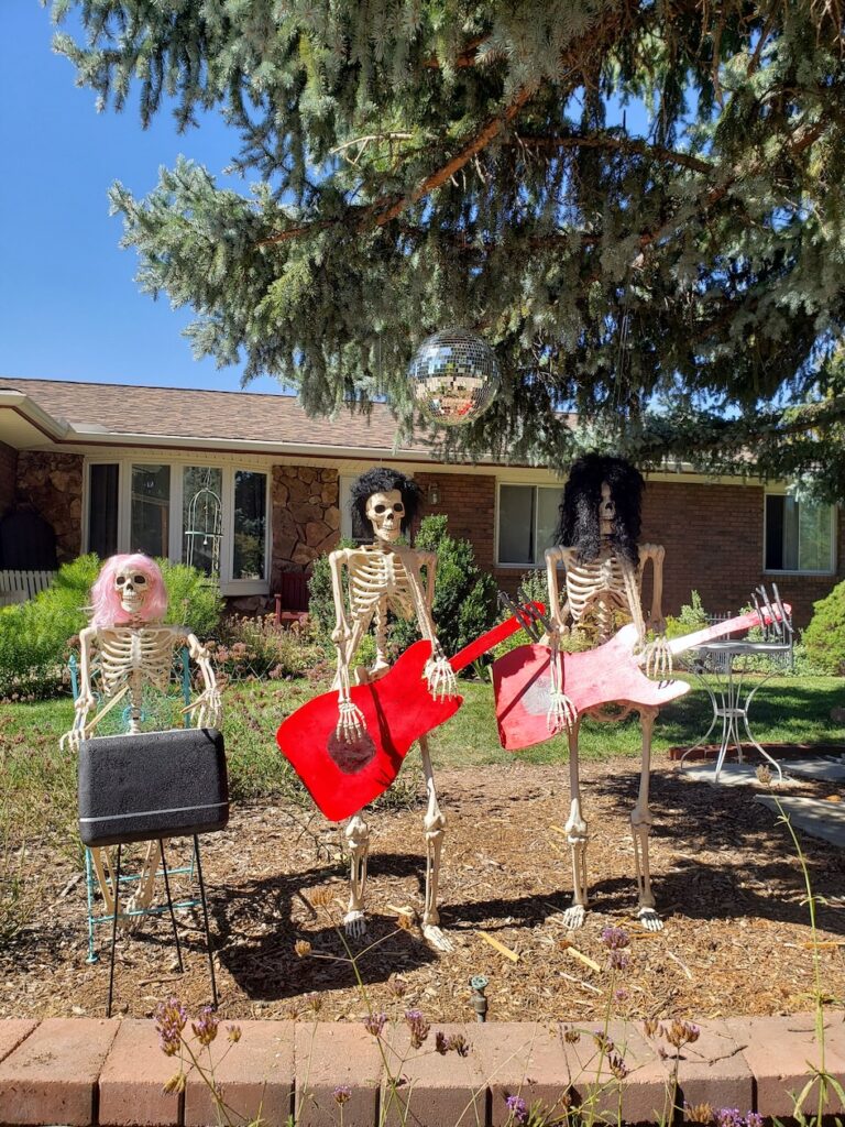 Three skeleton statues in front of a house wearinf wigs and playing guitars and pianos.