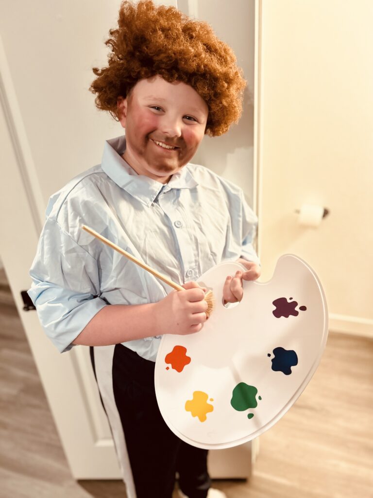 A school age boy dressed as Bob Ross with vibrant red hair. He smiles while holding a paint palette filled with various colors and and a brush.